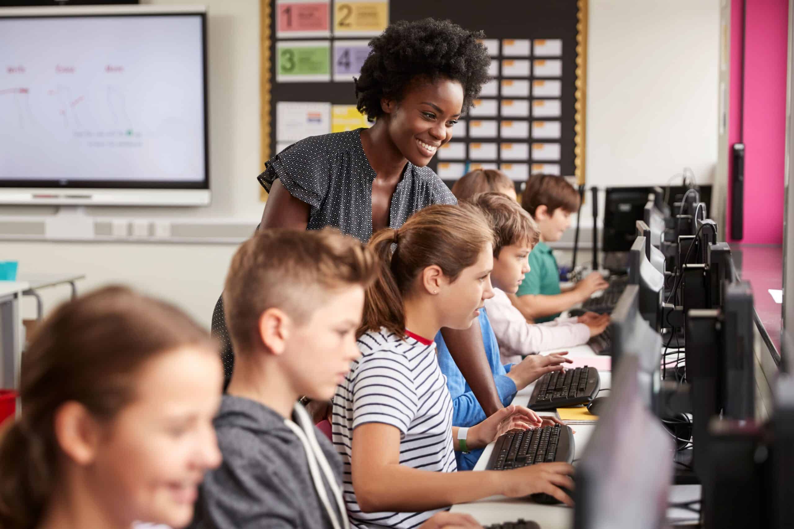A smiling teacher guides students working on computers in a classroom, reflecting the dynamic and engaging environment of Secondary Teacher jobs in Peterborough