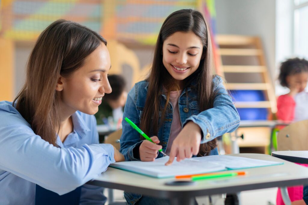 A teacher guiding a student with a smile in a classroom, reflecting the supportive roles provided by teaching recruitment services in Peterborough